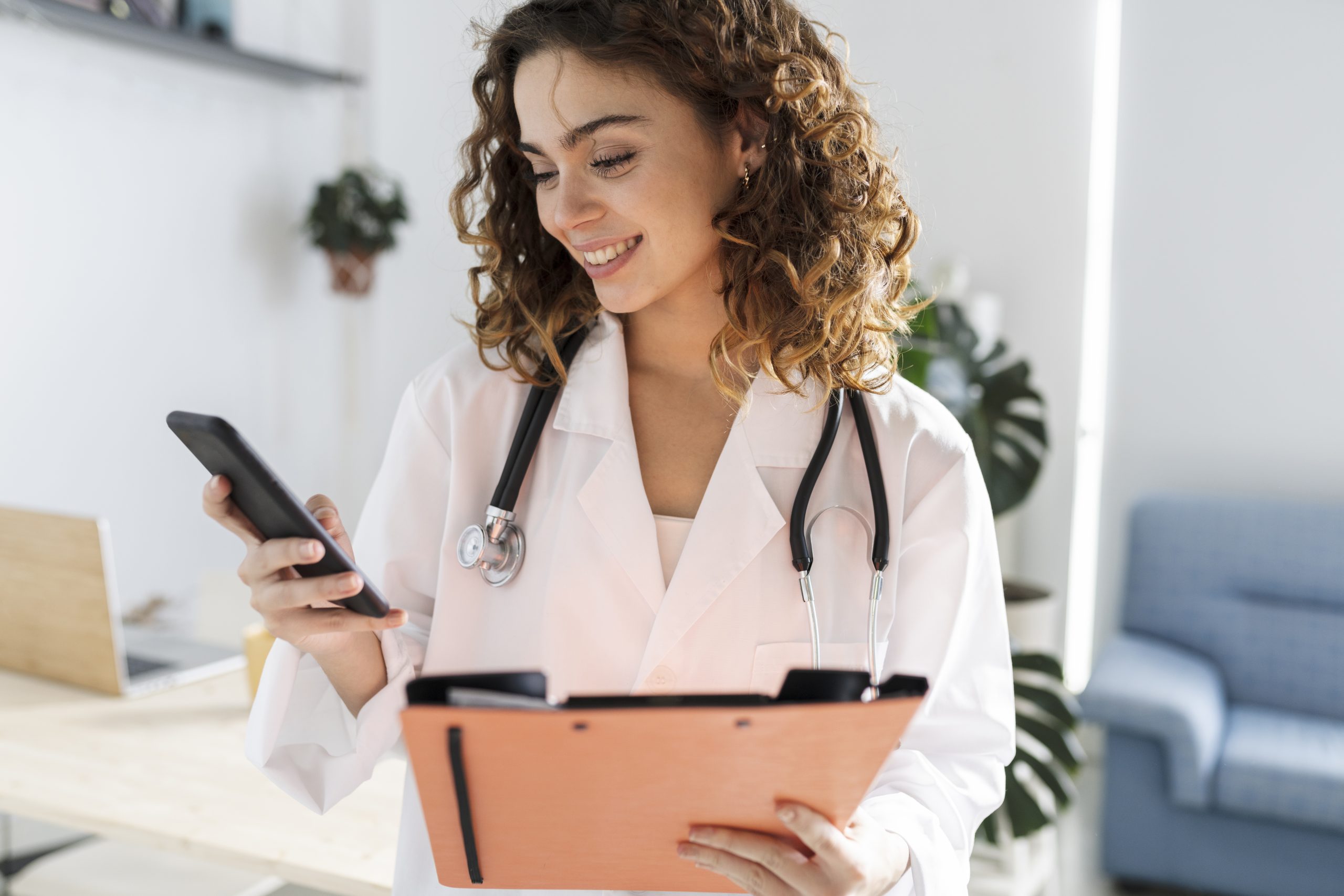 Un médecin en blouse blanche avec un stéthoscope vérifie son téléphone tout en tenant un presse-papiers dans un bureau.